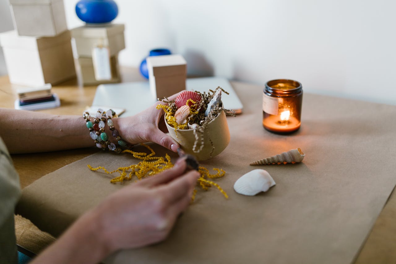 Hand arranging seashells on craft paper with a lit scented candle in calm, creative setting.