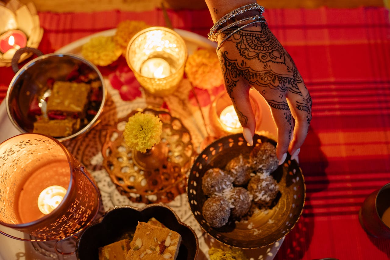 A hand with henna design reaches for sweets among Diwali candles and decorations.