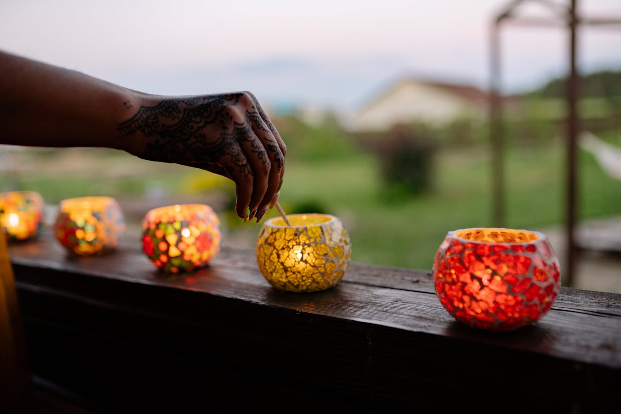 services-04 A henna-tattooed hand lighting colorful glass candles on a wooden surface at twilight.